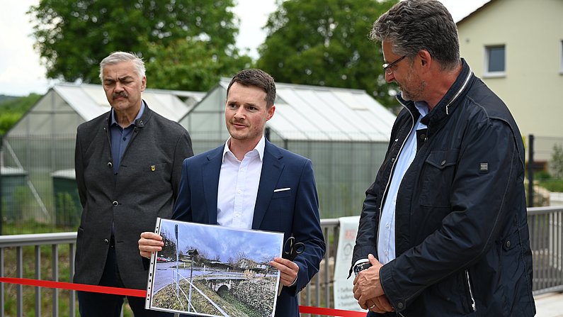 Auf dem Bild sind drei Männer zu sehen. Einer hält ein Foto in der Hand. 