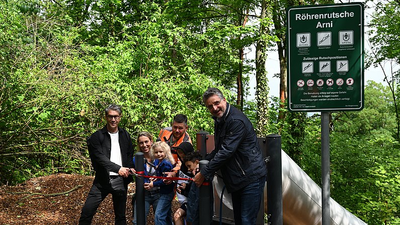 AUf dem Bild sind Kinder und Erwachsene zu sehen. Sie schneiden vor einer Rutsche ein rotes Band durch. 