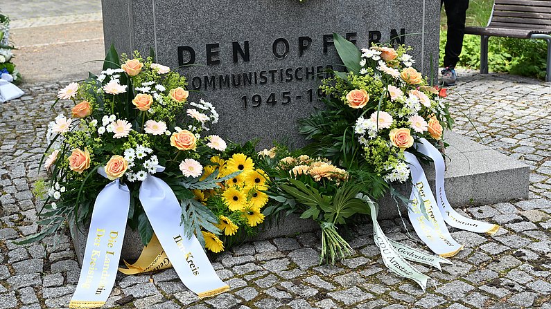 Auf dem Foto sieht man Blumengebinde an einem Gedenkstein stehen. 